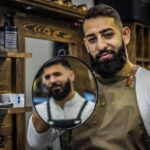 Beard Seborrheic Dermatitis: Causes, Symptoms, and Treatments - A bearded barber smiling while holding a mirror in a stylish barbershop. Ideal for lifestyle and grooming themes.
