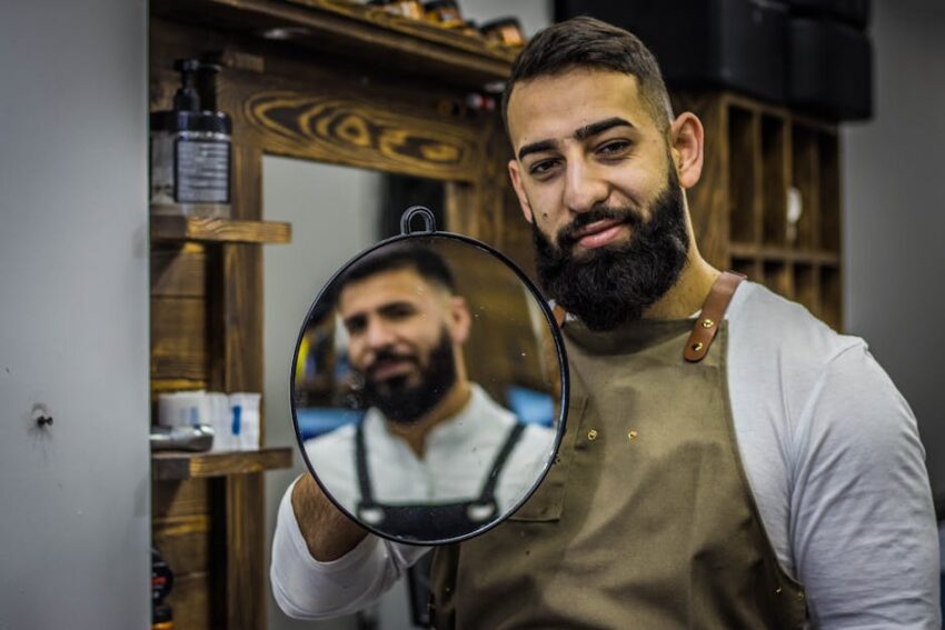 Beard Seborrheic Dermatitis: Causes, Symptoms, and Treatments 24 Beard Seborrheic Dermatitis: Causes, Symptoms, and Treatments - A bearded barber smiling while holding a mirror in a stylish barbershop. Ideal for lifestyle and grooming themes.