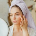 Best Face Moisturizers for Seborrheic Dermatitis 2023: Top 16 Picks - Woman with head towel applying moisturizer in front of mirror indoors.