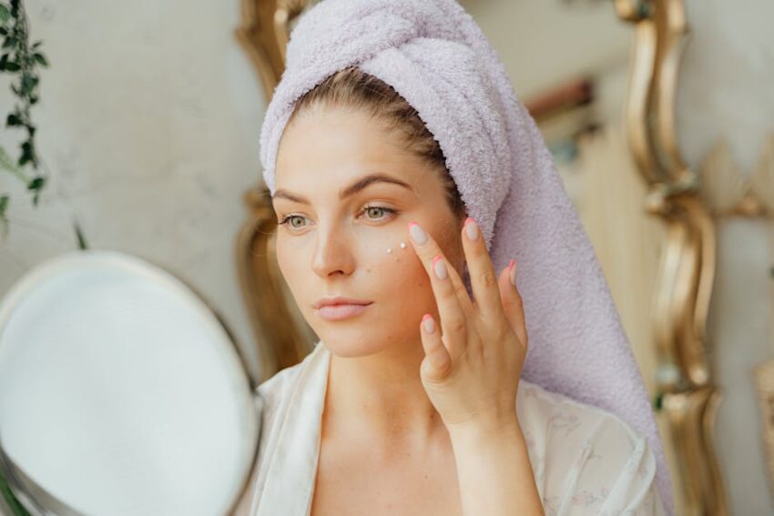 Best Face Moisturizers for Seborrheic Dermatitis 2023: Top 16 Picks - Woman with head towel applying moisturizer in front of mirror indoors.