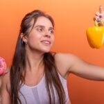 Seborrheic Dermatitis Diet Guide: Foods That May Help and What to Avoid (2026) - Woman in sports bra juggling choice between a donut and bell pepper on orange background.