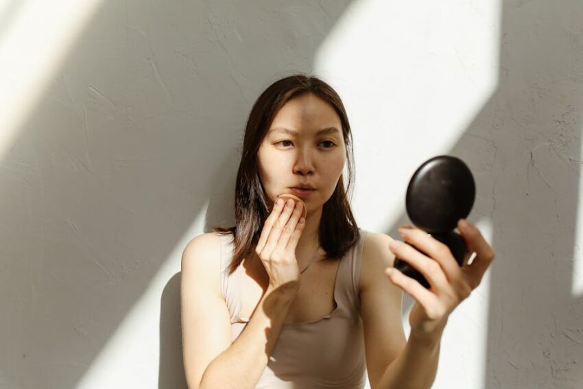 best-sunscreen-seborrheic-dermatitis-2026 - Young Asian woman applying makeup with a mirror in soft natural lighting indoors.