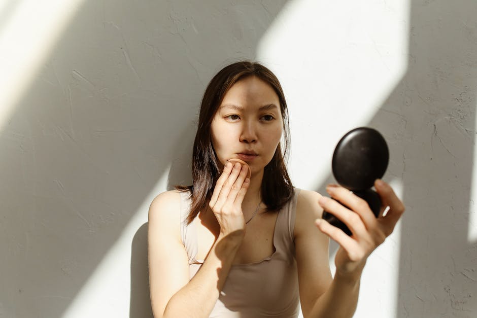 best-sunscreen-seborrheic-dermatitis-2026 - Young Asian woman applying makeup with a mirror in soft natural lighting indoors.