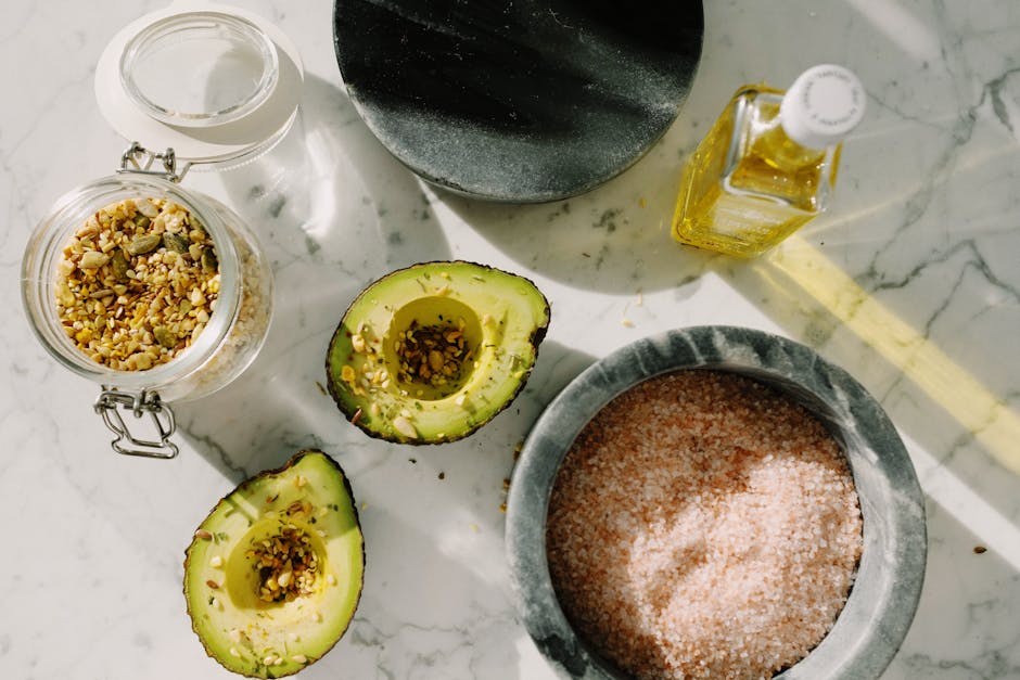 Can Diet Affect Seborrheic Dermatitis? What 2026 Research Shows - Top view of tasty halved avocado sprinkled with various seeds placed near pink salt with seed mixture in glass pot and other dressing ingredients arranged on white marble table