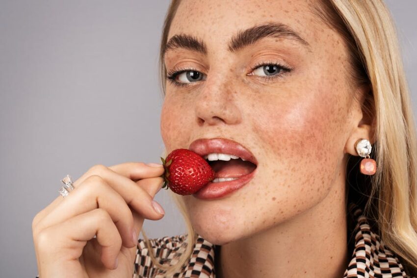 malassezia-safe-diet-seborrheic-dermatitis - Close-up portrait of a blonde woman holding a strawberry near her lips.