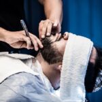 tea-tree-oil-seborrheic-dermatitis-guide-2026 - An adult man receiving a precise beard trim in a barbershop with a warm towel treatment.